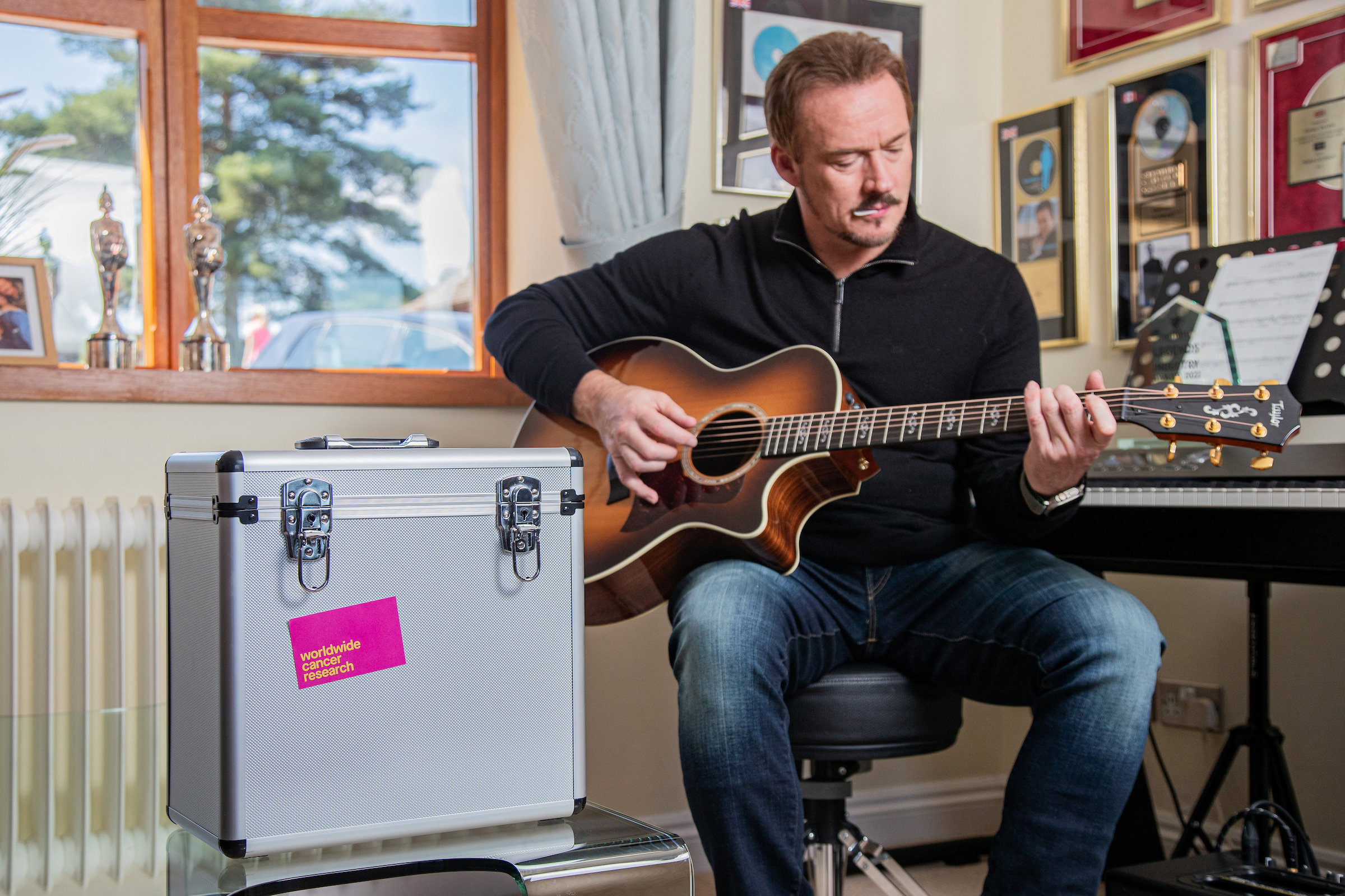 Singer Russell Watson playing guitar next to a silver record case with the Worldwide Cancer Research logo on